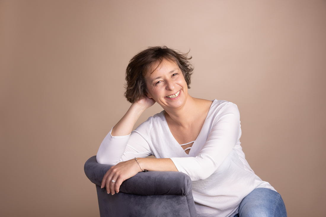 femme souriante assise sur une chaise, vêtue d'un haut blanc et d'un jean, avec un fond beige.